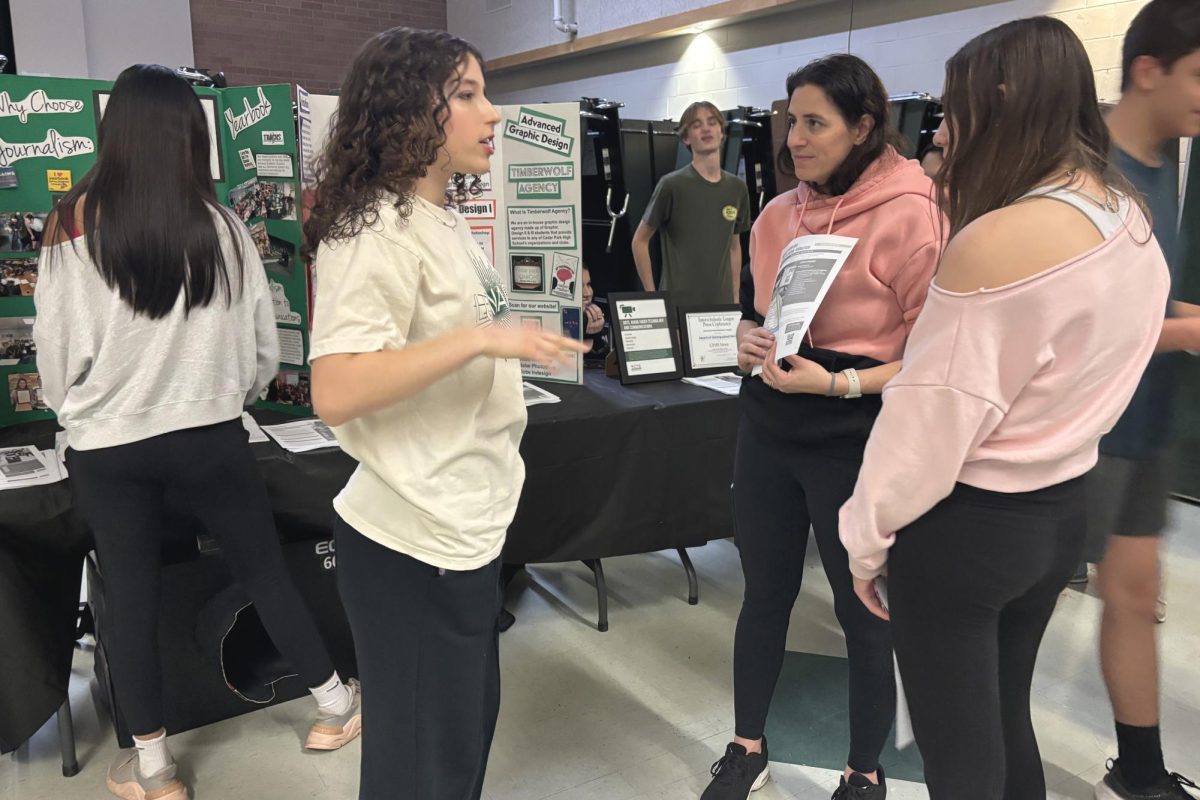 Standing in front of the Digital Communications booth, junior Ava Holmes discusses the Graphic Design classes with eighth grade parents and their students during CP night on Jan. 28. Holmes is a member of Timberwolf Agency, the advanced graphic design class, and is working towards her Adobe Certification. "It's important to be certified and show them that you're capable of all the different tasks in Illustrator," Holmes said. "Repetition is key to passing. Just do it again and again, and if you don't get it right, do it again." Photo by Alyssa Fox